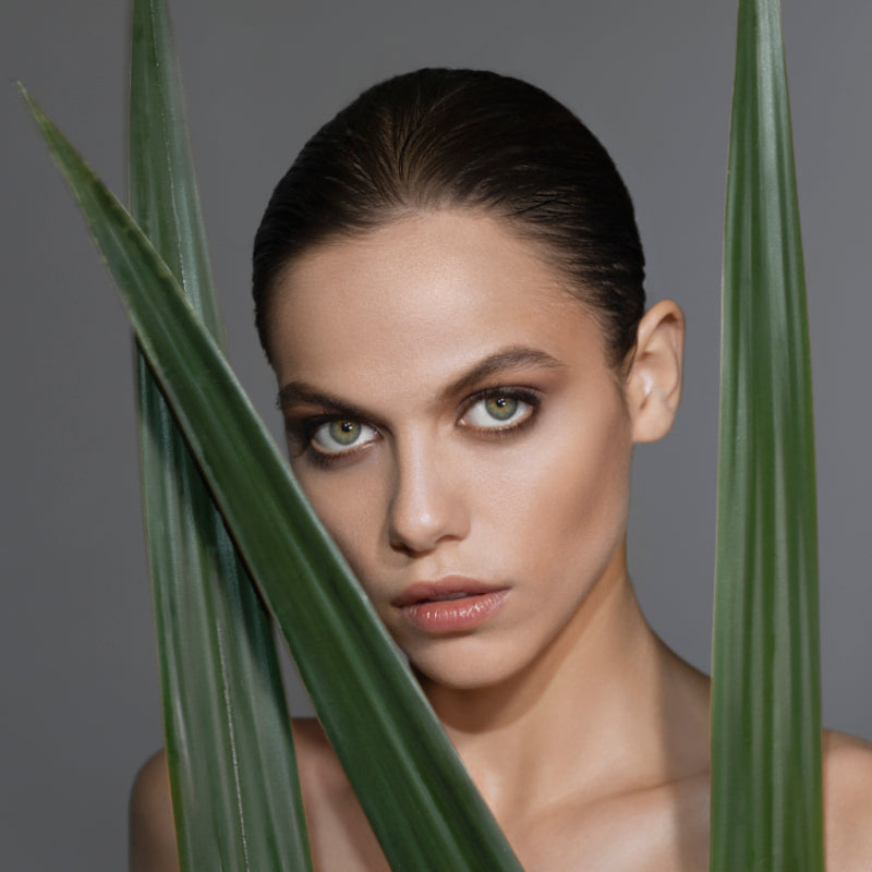 Woman with green eyes and hair pulled back, framed by large green leaves against a gray background